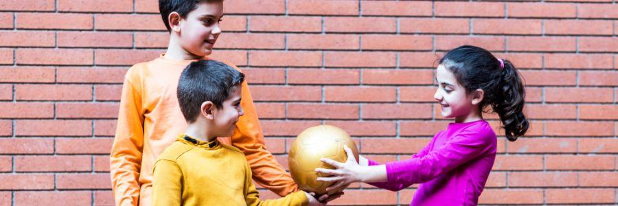 Three children sharing a ball while smiling, symbolizing friendship and inclusion — a simple act that reflects day of giving ideas during school recess.