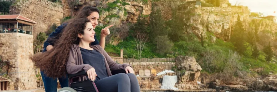 Two young women enjoy a scenic view, one in a wheelchair and the other pointing ahead, symbolizing freedom, inclusion, and the spirit of National Disability Independence Day.