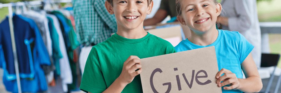 Two smiling children holding a cardboard sign that says “Give” while participating in a community donation activity. 