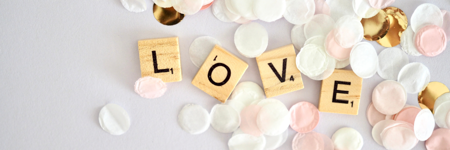 Scrabble-style tiles spelling LOVE surrounded by soft pink, white, and gold confetti 