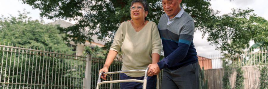 Older man helping an older woman use a walker outdoors, showing care, support, and daily giving 