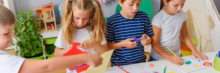 Four children sit together at a table engaged in a hands-on craft activity, cutting and drawing colourful paper shapes in a bright, cheerful classroom setting.