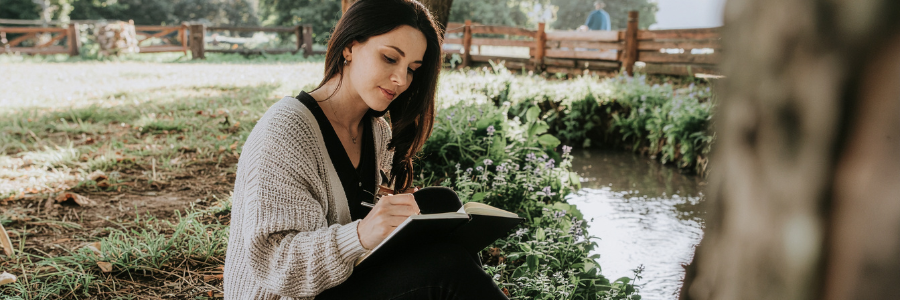 Woman journaling beside a peaceful stream as part of her daily self-care routine.