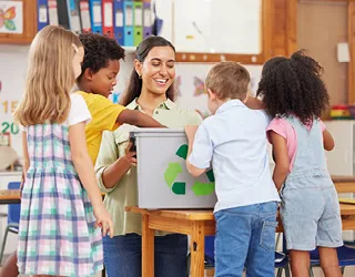 A smiling teacher with diverse young students gathered around a recycling bin in a classroom, fostering a happy and thriving community as part of the 365give Challenge.