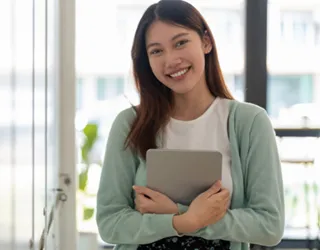 A young woman standing by a bright window, smiling while holding a tablet, ready to make a difference.