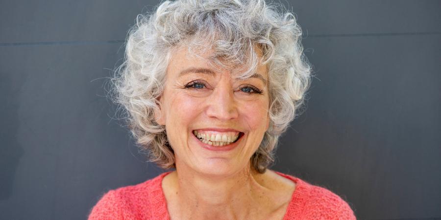 Smiling older woman with curly gray hair wearing a coral top against a dark background.