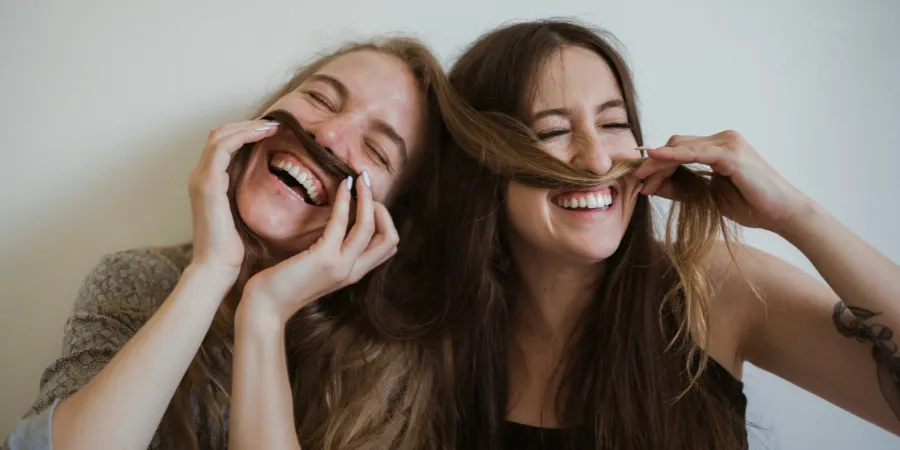 Two women laughing and smiling together while playfully holding strands of hair across their faces.
